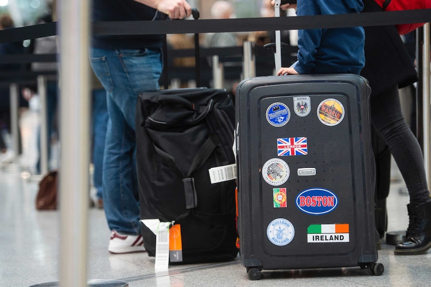 Two adults carry baggage behind airport queue, only their hands and legs are visible, as a child sits on one of the suitcases.