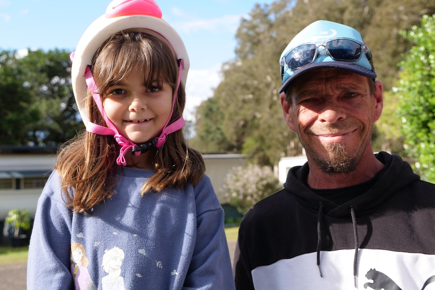 Father and seven year old daughter smiling in the sun at a caravan park, she wears a bike helmet.