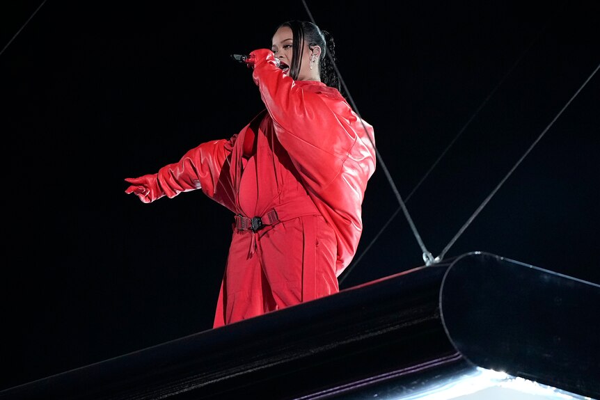 Rihanna with mic up and hand out wearing a red jumpsuit