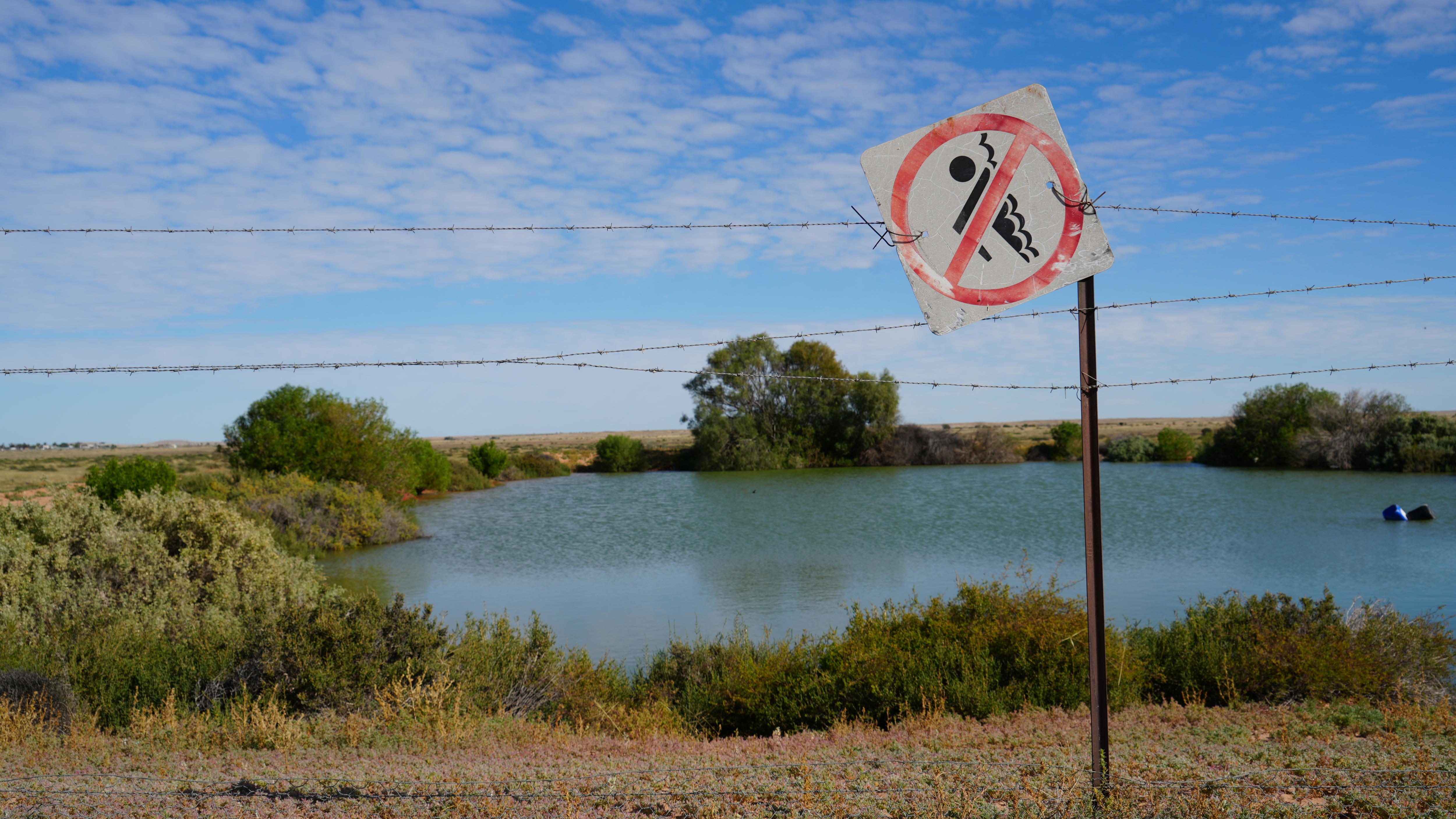 A 'no swimming' sign on a wire fence infront of a dam full of water