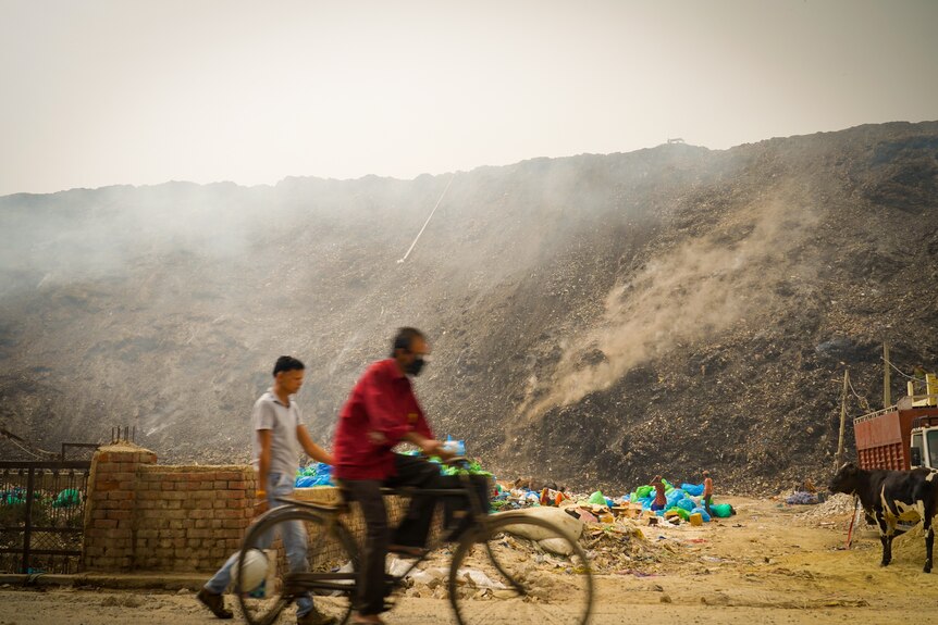 A man cycles past a massive pile of smouldering garbage 