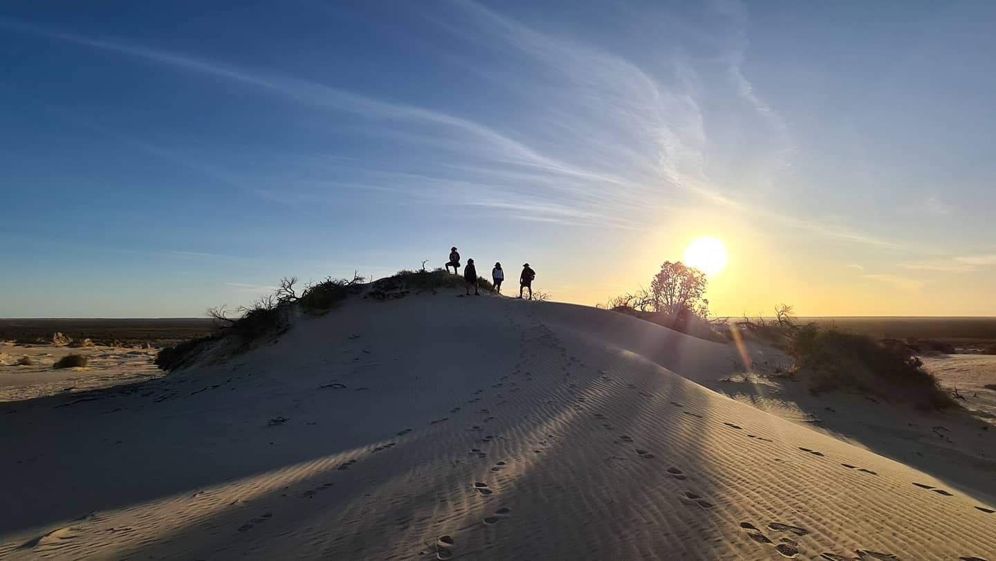 Footprints lead to four people who stand at a distance on a small white sand hill as the sun sets behind them.