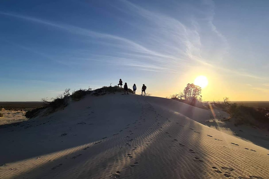 Footprints lead to four people who stand at a distance on a small white sand hill as the sun sets behind them.