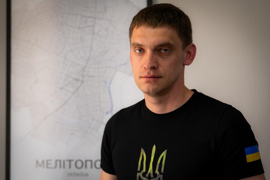 A young man wearing a black Tshirt with Ukraine flag patch on one sleeve stands in front of a map of Melitopol