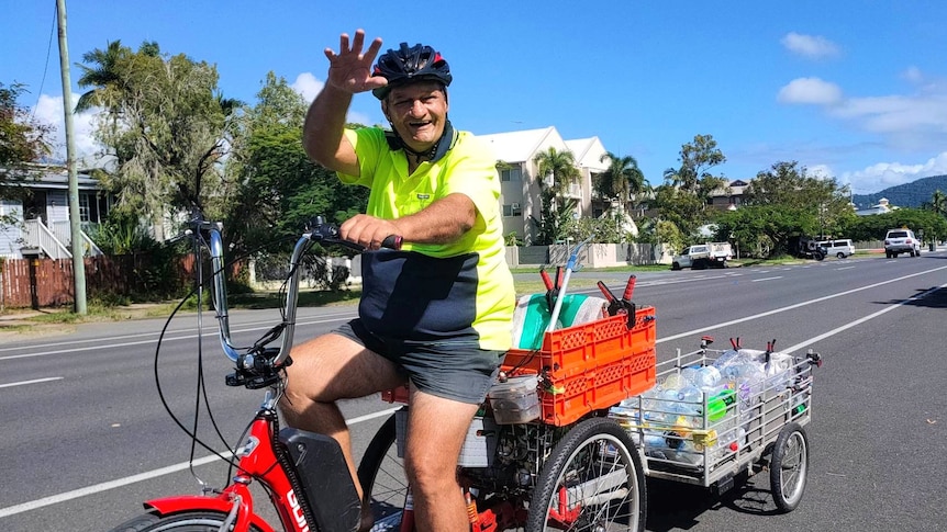 A man in a high-vis shirt and helmet waves while riding a red tricycle pulling a trailer full of used cans and bottles