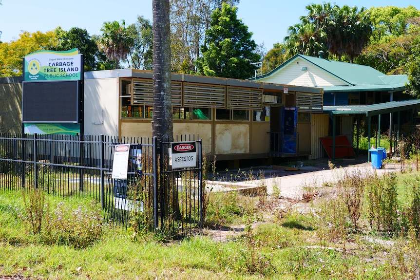 A small school building with a danger sign hanging on its fence. It is surrounded by weeds.