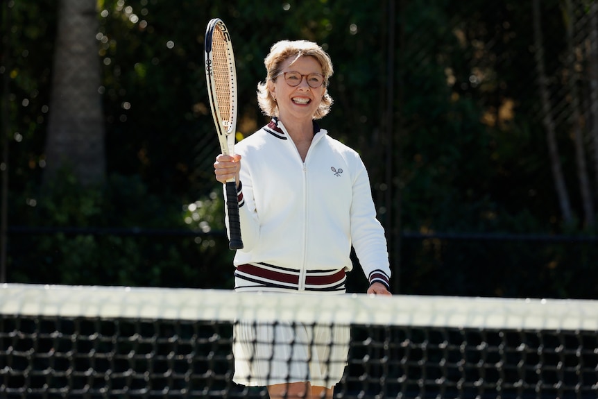 A TV still of Annette Bening on a tennis court. She's smiling, and holding her racket up.