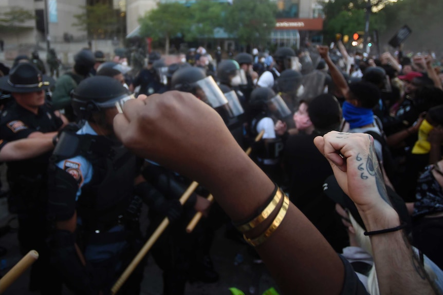 Two fists raised in the air with riot police advancing in the background.