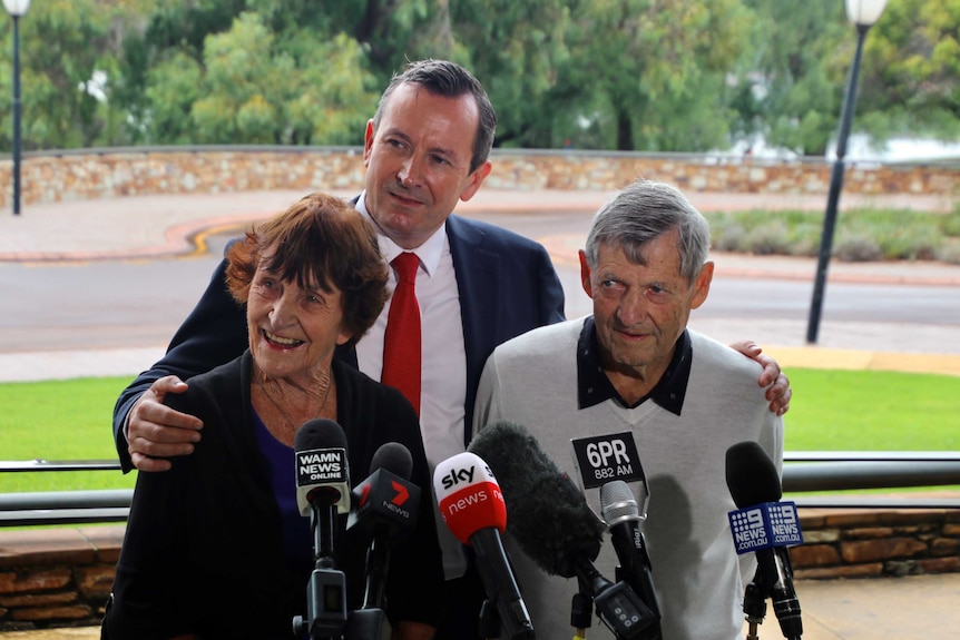 WA Premier Mark McGowan poses for a photo standing behind his parents with microphones in front of them.