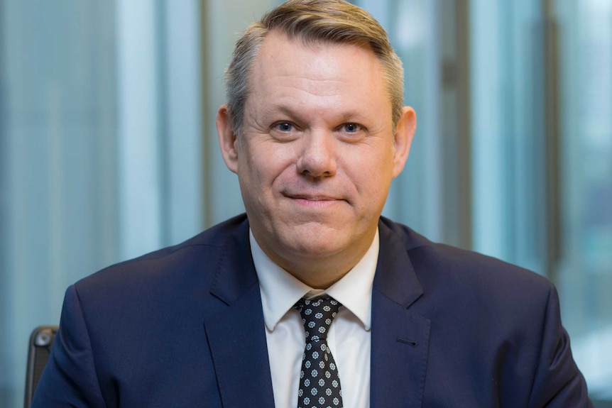 A man with short, blond hair, wearing a dark suit and sitting in an office.