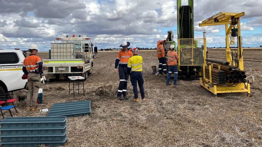 Workers stand near survey equipment in a bare paddock.