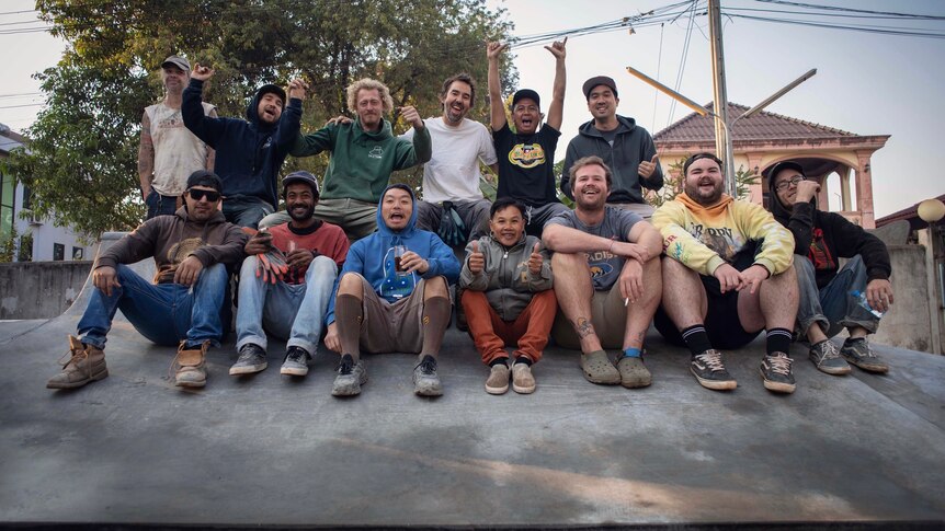 A group of men of varying ethnicities and outfits smiling and sitting on a concrete skate ramp.
