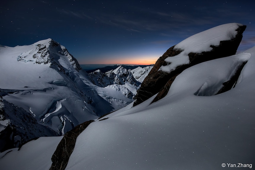 Sun peaks behind snow covered mountain tops. Dark skies surrounds. 