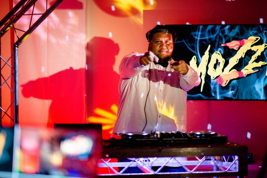 A man wears a shirt and has headphones adopt his head. He stands at a DJ's deck and points two fingers to camera.