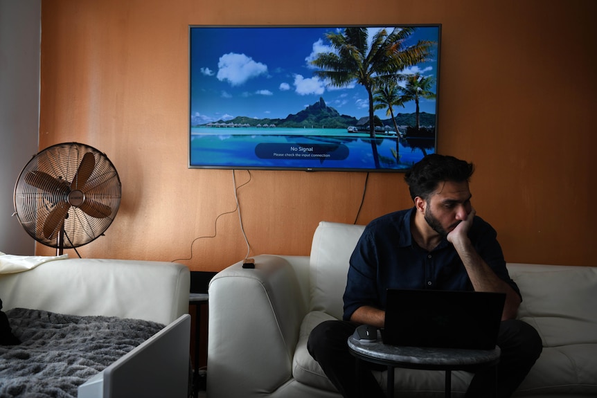 A 26-year-old man with dark hard and trimmed beard sits on a white counch looking down on his laptop that sits on a coffee table