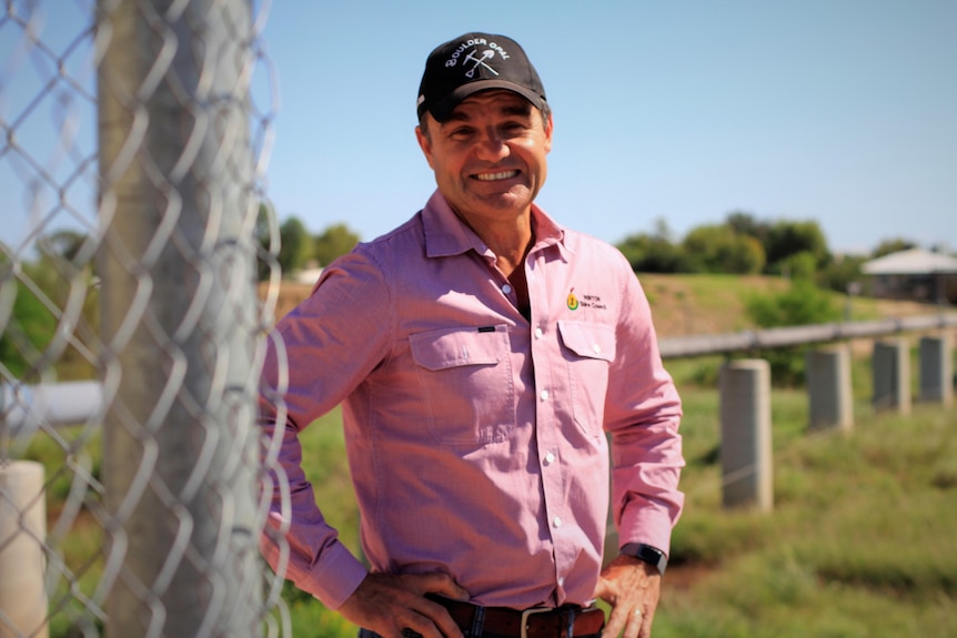 A smiling middle-aged man in a dark cap and a button-up shirt stands in the sun with his hands on his hips.