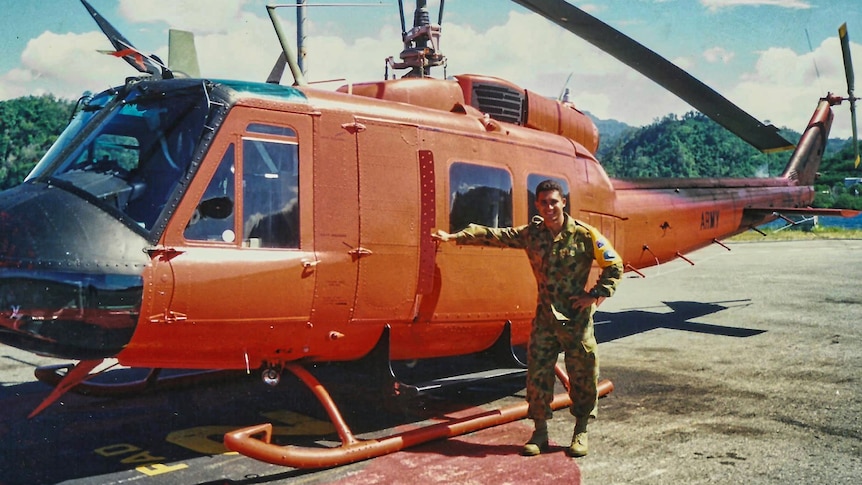 A man in camouflage uniform smiling and standing by a orange helicopter.
