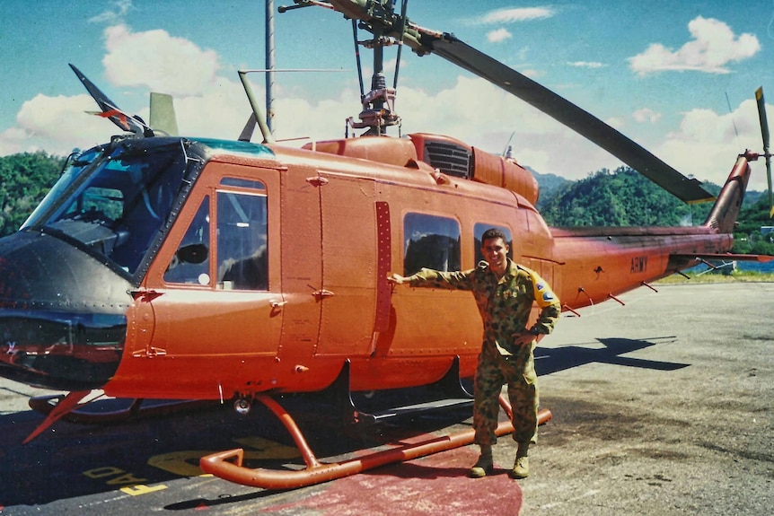 A man in camouflage uniform smiling and standing by a orange helicopter.