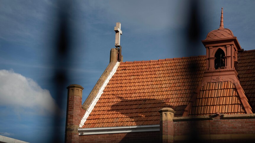 A view of a church roof, with a white cross at the top, seen through out-of-focus black bars of a fence