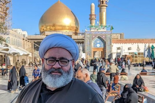 A photo of Sheikh Mohammed Mehdi at the shrine in Karbala. The gold dome is shown, with people around him in congregation.