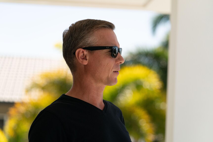 A man in black sunglasses looks into the distance with a palm tree in the background