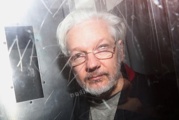 Julian Assange regarde la caméra alors qu'il est photographié derrière une vitre avec des graffitis gravés dessus.  Ses cheveux gris sont de retour.