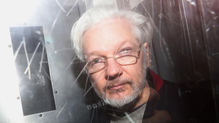 Julian Assange looks to the camera as he is photographed from behind glass with graffiti etched into it. His grey hair is back.