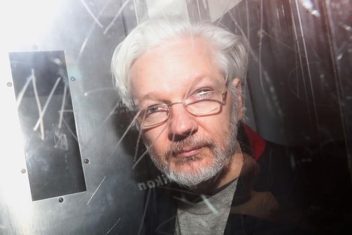 Julian Assange looks to the camera as he is photographed from behind glass with graffiti etched into it. His grey hair is back.