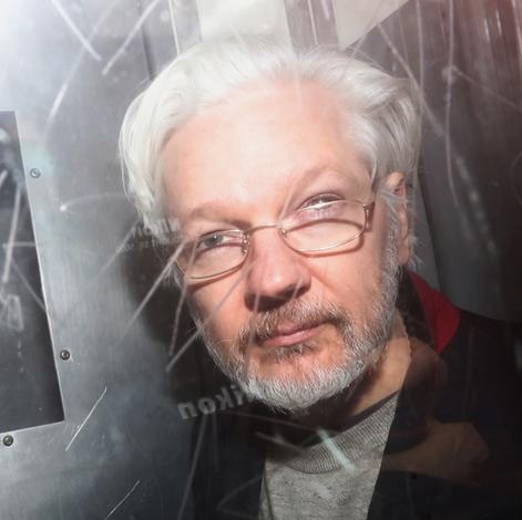 Julian Assange looks to the camera as he is photographed from behind glass with graffiti etched into it. His grey hair is back.