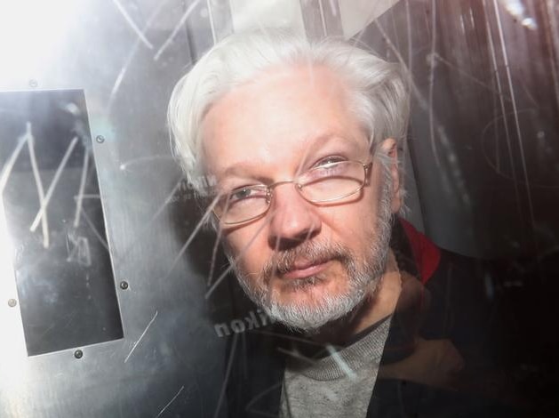 Julian Assange looks to the camera as he is photographed from behind glass with graffiti etched into it. His grey hair is back.