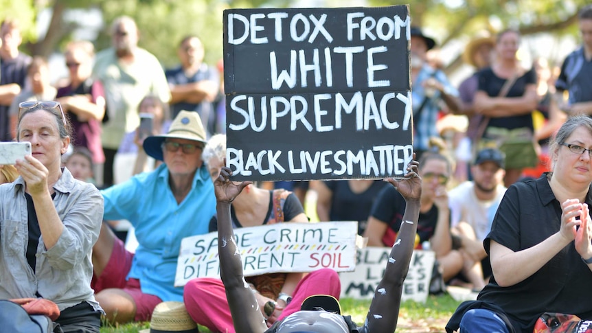 A man is lying on the ground holding up a sign which says 'detox from white supremacy; black lives matter'
