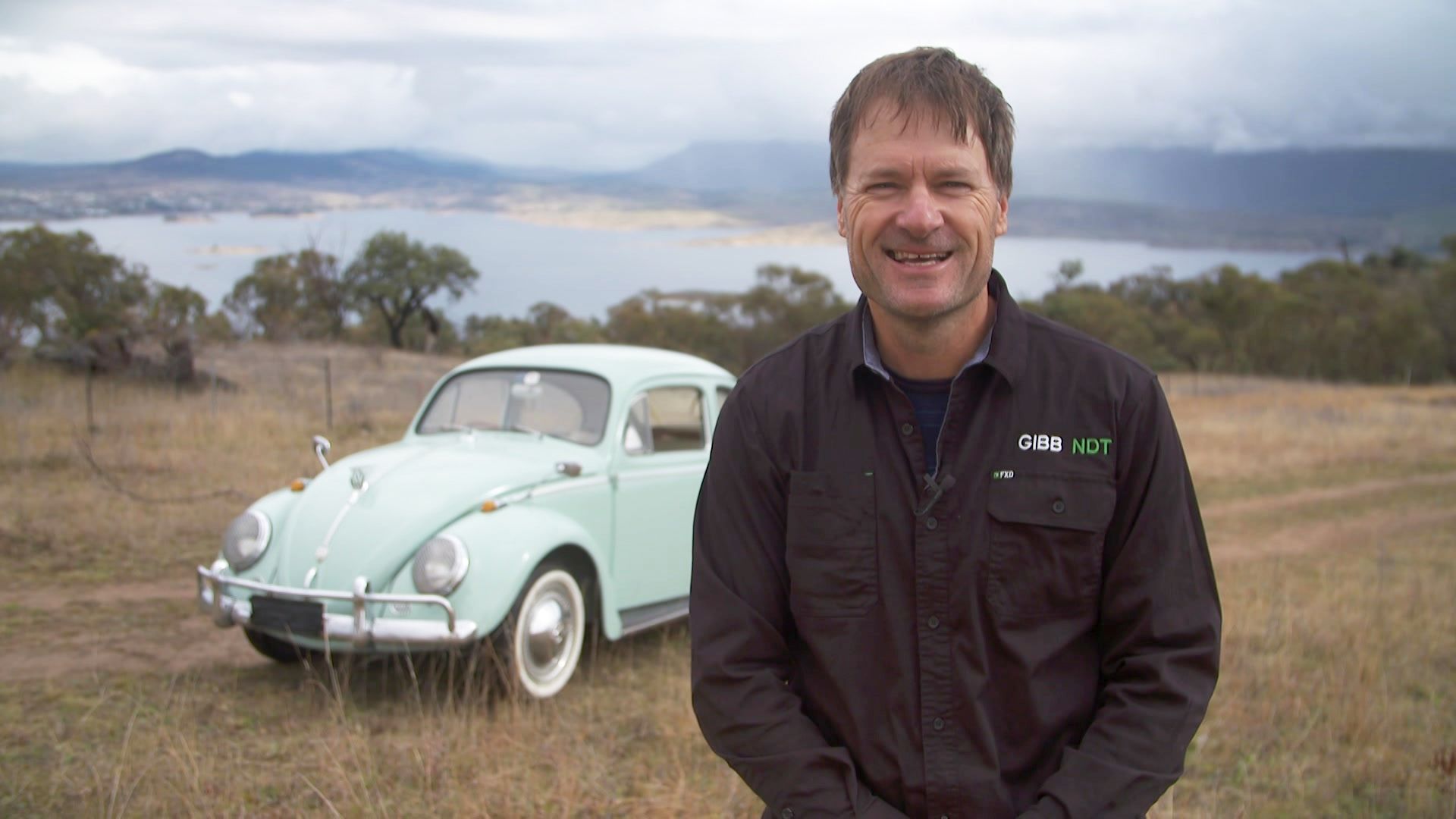 Conrad stands in front of his blue VW beetle in a park.