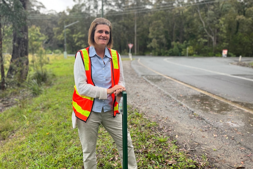 Woman in high-vis orange vest standing next to green fence post on side of road.,