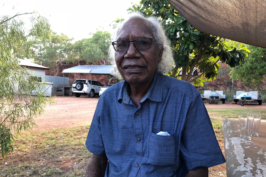 Indigenous man Ross Williams sits on a park bench, next to cars and trailers.