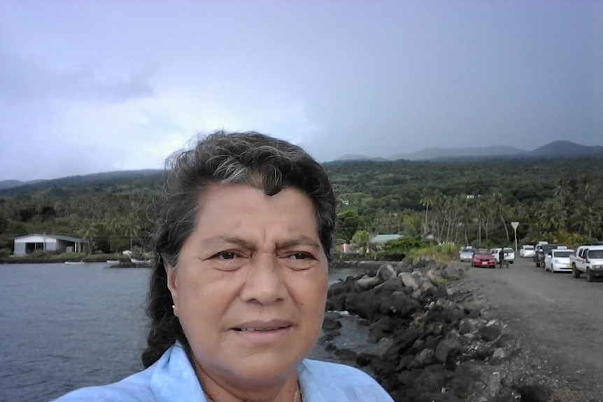 A woman wearing a light blue button up shirt taking a selfie in front of an island in Fiji.