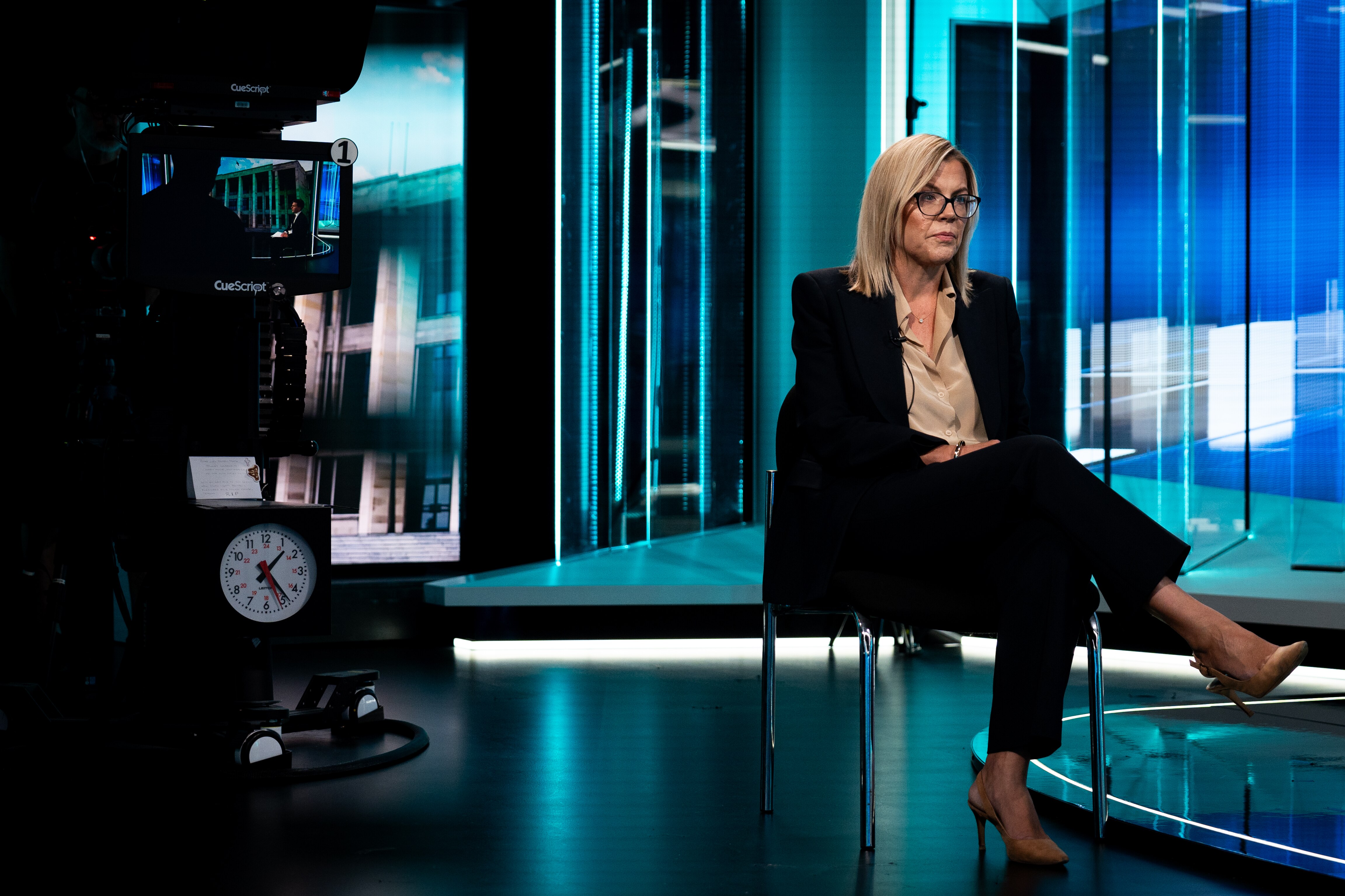 A wide shot of Libby Mettam in a dark pantsuit sitting in a television studio, surrounded by camera equipment.