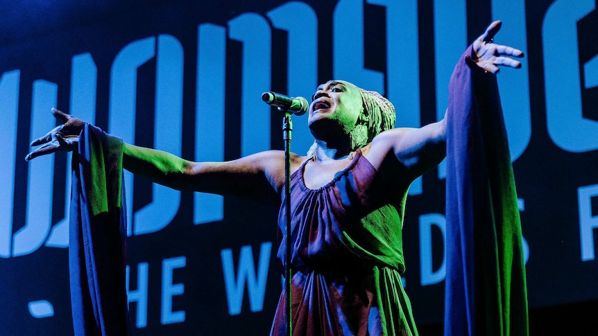Zaachariaha Fielding sings into a microphone with their arms outstretched in front of a screen displaying the word 'WOMADELAIDE'