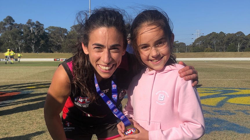 A young girl dressed in pink holds a premiership medallion next to an AFLW player.
