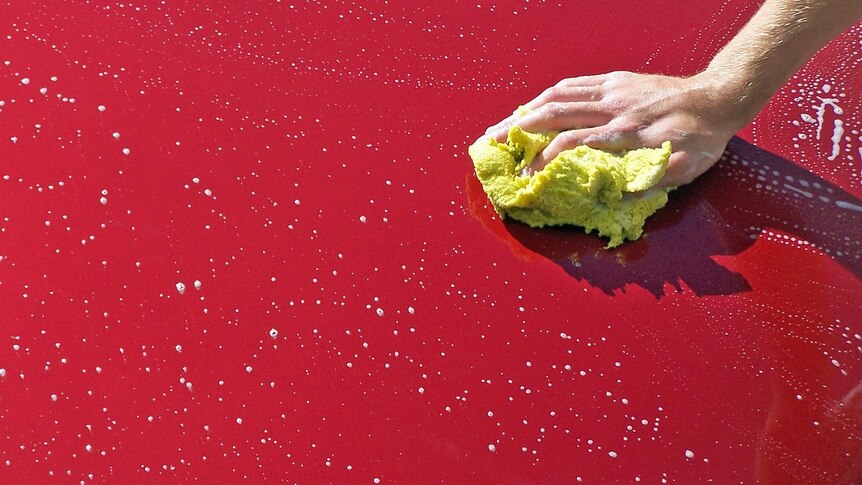 A hand with a sponge rests on a red car bonnet covered in suds.
