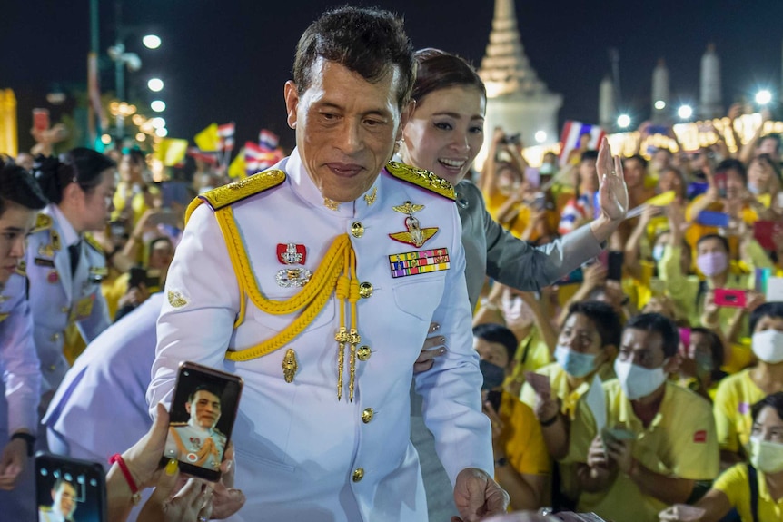 Thailand's King smiles for crowds taking pictures on their phones as queen waves in background.