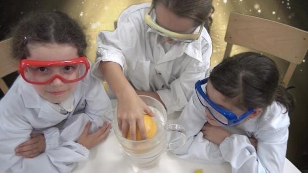 Children in white coats and safety glasses stand with a floating grapefruit in a jug of water