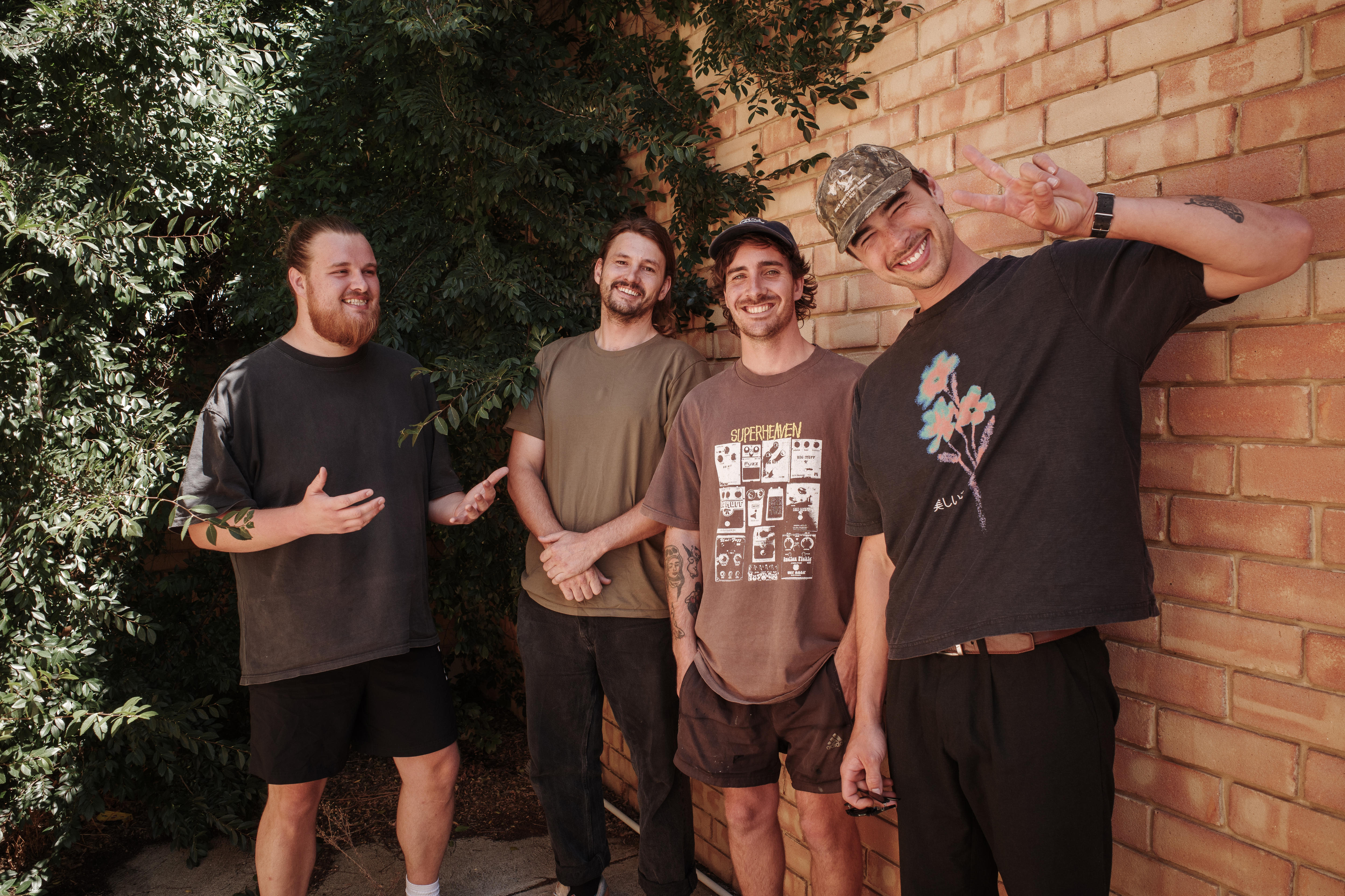 Four men stand together smiling and posing in front of a sandstone wall and large bush