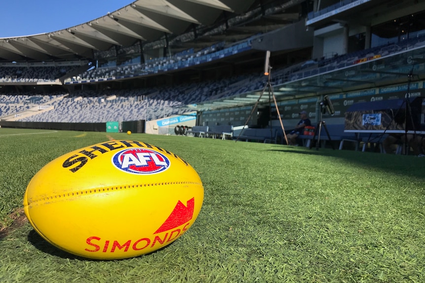 A yellow football lies on the grass of a football stadium, and in the background there is an empty stadium and the blue sky.