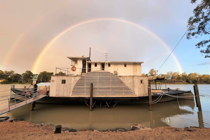 A steam boat sits on a river bank, with a rainbow in the background