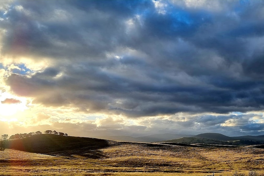 A stunning landscape photo of hills in the Snowy Monaro, with a cloudy sky and sun poking through