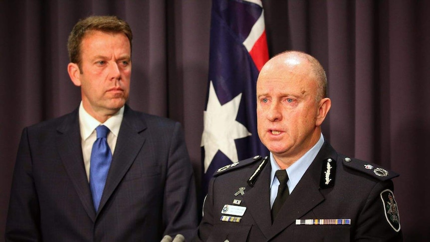 Dan Tehan watches as Neil Gaughan, in his AFP uniform, speaks at a lectern. There is an Australian flag in the background.