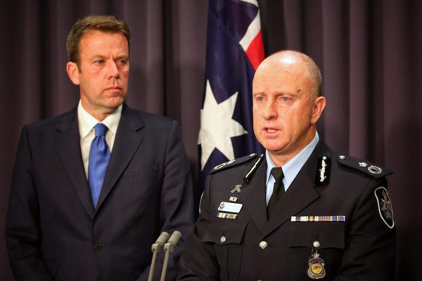 Dan Tehan watches as Neil Gaughan, in his AFP uniform, speaks at a lectern. There is an Australian flag in the background.