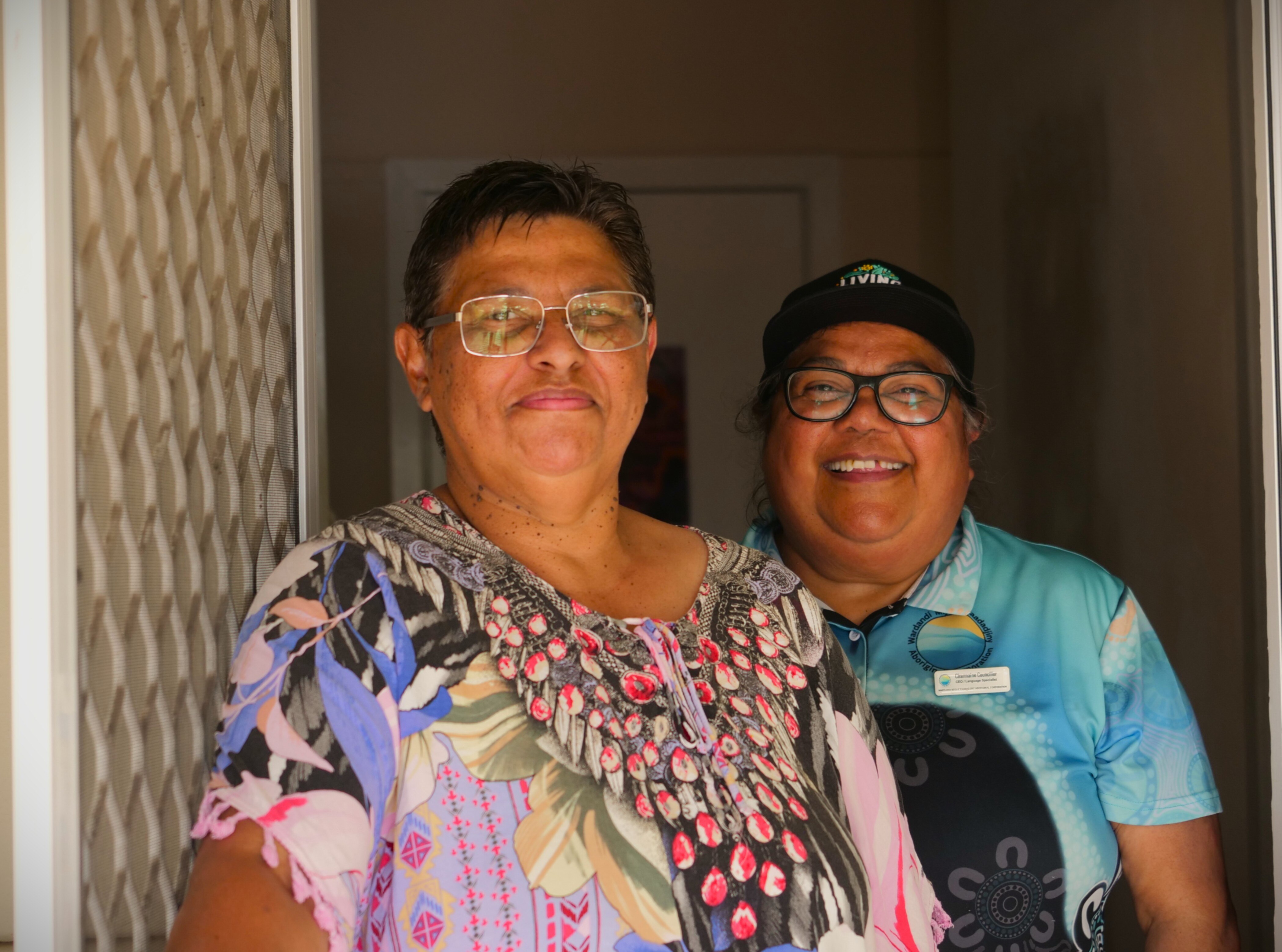 Dos mujeres están paradas en una puerta, sonriendo a la cámara y sosteniendo una puerta de seguridad abierta.