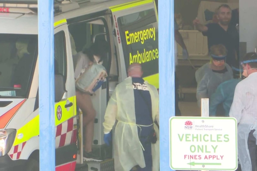 A woman with a face mask on gets out of an ambulance as medical staff surround her