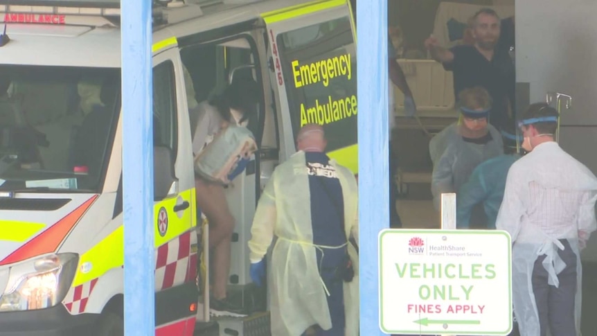 A woman with a face mask on gets out of an ambulance as medical staff surround her